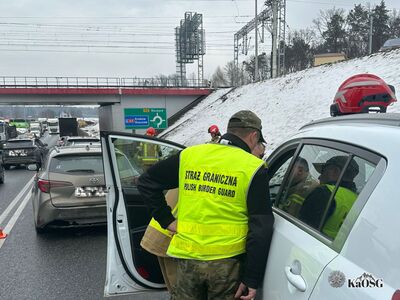 Akcja ratunkowa na drodze ekspresowej w zimowej scenerii. Na pierwszym planie funkcjonariusz Straży Granicznej w odblaskowej żółtej kamizelce oraz strażak pochylają się nad białym samochodem osobowym z otwartymi drzwiami. Nad drogą przebiega wiadukt kolejowy, a obok znajduje się zaśnieżona skarpa. Na zielonej tablicy drogowej widnieje kierunek na Kraków i Rzeszów (autostrada A4).