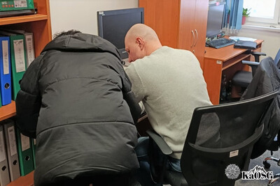 Zatrzymany cudzoziemiec podpisuje na biurku dokumenty. Towarzyszy mu funkcjonariusz w stroju cywilnym.