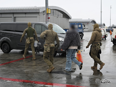 Mężczyźni w zielonych mundurach, z kominiarkami na głowach prowadzą do samochodu służbowego kobietę Kobieta jest ubrana w dżinsy, szare futerko  a w dłoni trzyma pomarańczową siatkę.