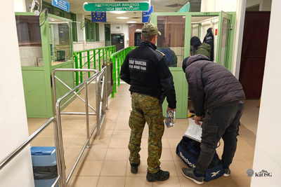 Na zdjęciu mężczyzna po prawej stronie ubrany jest w mundur, drugi schyla si e do torby podróżnej która leży na podłodze. Mężczyźni stoją na hali odprawy granicznej na przejściu granicznym.