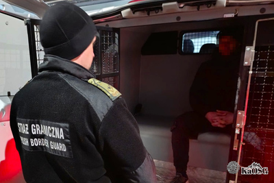 Na zdjęciu widać funkcjonariusza Straży Granicznej stojącego tyłem. Patrzy na mężczyznę, który siedzi w samochodzie specjalnym do przewożenia osób zatrzymanych