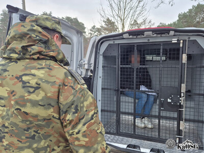 zatrzymany cudzoziemiec siedzi w pojeździe służbowym sg typu więźniarka, asystuje mu funkcjonariusz