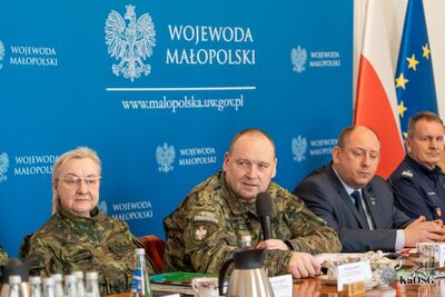 Funkcjonariusz Straży Granicznej w mundurze polowym przemawia do mikrofonu podczas obrad. Obok niego siedzą inni uczestnicy w strojach cywilnych i mundurowych na tle niebieskiej ścianki z napisem „Wojewoda Małopolski”.