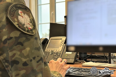 przy biurku siedzi funkcjonariusz sg, widać jego ramię i dłoń przy klawiaturze komputera. na biurku stoi monitor, w tle po lewej stronie okno