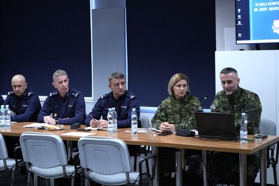 Pięć osób siedzi przy stole na konferencji , trzy z nich po lewej stronie ubrani są w granatowe mundury, dwie od strony prawej mają mundury zielone.