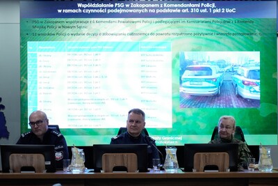 Na zdjęciu widać trzy osoby siedzące przy stole konferencyjnym. W tle widać wielki monitor, na którym wyświetlane są informacje.