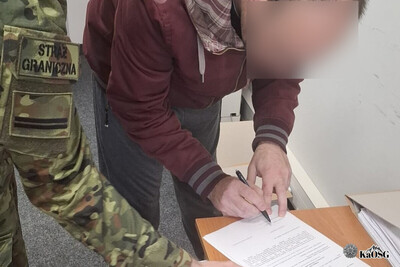Mężczyzna podpisuje dokument w pozycji schylonej. Dokument leży na stole. Z lewej strony widoczny fragment ręki mężczyzny ubranego w mundur z napisem Straż Graniczna.