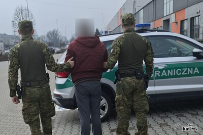 Dwaj mężczyźni ubrani w mundury moro, na głowach czapki z daszkiem, do pasa przypięta broń, prowadzę pod rękę trzeciego mężczyznę ubranego w cywilne ubranie, najprawdopodobniej jest to osoba zatrzymana. W tle samochód służbowy z kogutami i napisem Straż Graniczna.