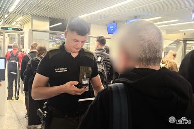 Na zdjęciu funkcjonariusz Straży Granicznej na terenie portu lotniczego kontroluje dokumenty. Przed nim stojący tyłem pasażer. W tle hala odlotów lotniska.