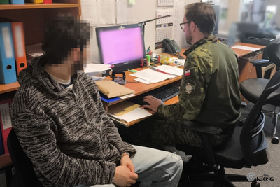 Na zdjęciu widać dwie osoby, które siedzą. Jeden siedzi na wprost komputera ubrany jest w mundur.