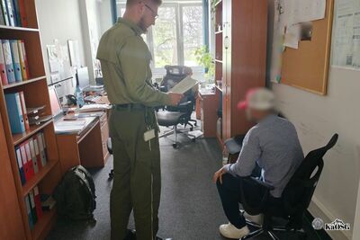 Na zdjęciu pomieszczenie biurowe. Po prawej stronie tablica korkowe i wiszące na niej dokumenty. Po stronie lewej szafa biurowa z segregatorami w różnych kolorach. Po środku stoi mężczyzna, prawdopodobnie funkcjonariusz ubrany w mundur (koszula i spodnie typu bojówki)  , trzyma w rękach dokumentu. Tuż obok niego siedzi inny mężczyzna, w cywilnym ubraniu, prawdopodobnie jest to osoba zatrzymana.