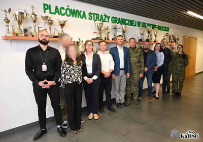 uczestnicy spotkania stoją do pamiątkowego zdjęcia na tle ściany, na której znajdują się półki z pucharami i napis Placówka straży granicznej w krakowie-balicach