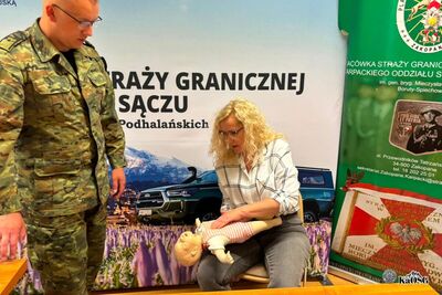 Na zdjęciu, po lewej stronie funkcjonariusz Straży granicznej w mundurze moro. Na ramieniu naszywka z napisem Straż Graniczna. Obok niego w pozycji schylonej kobieta a przed nią manekin służący do ćwiczeń z pierwszej pomocy przedmedycznej.