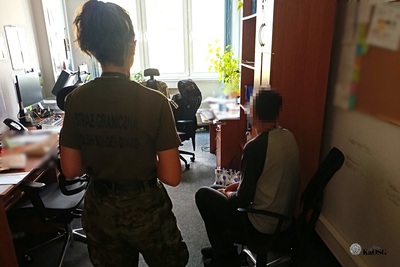 Na zdjęciu widać dwie osoby. Jedna siedzi, ma zamazaną twarz, kobieta w mundurze stoi po lewej stronie.