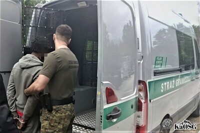 Na zdjęciu po prawej stronie mężczyzna w umundurowaniu  (spodnie moro i koszulka zielona z napisem na plecach Straż Graniczna) trzema pod rękę cudzoziemca, prawdopodobnie osobę zatrzymaną. Cudzoziemiec ma na rękach kajdanki i jest wprowadzony do samochodu służbowego typu więźniarka.