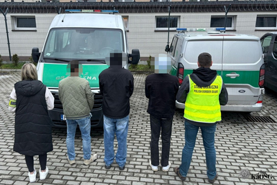 Na zdjęciu widać 5 osób, które stoją tyłem . Z przodu przed nimi stoją dwa samochody oznakowane w kolorze srebrnym z zielonymi napisami i pasami.