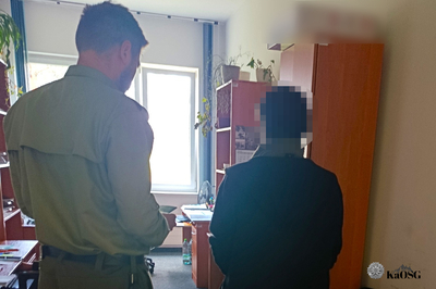 Na zdjęciu widać dwie osoby, które stoją tyłem w pomieszczeniu. Mężczyzna po lewej stronie jest ubrany w mundur i trzyma dokument w rękach.