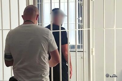 Na zdjęciu po lewej stronie mężczyzna w szarym T-shircie prowadzi pod rękę drugiego mężczyznę - prawdopodobnie zatrzymanego i prowadza go do pomieszczenia z kratami . Jest to prawdopodobnie pomieszczenie dla osób aresztowanych.