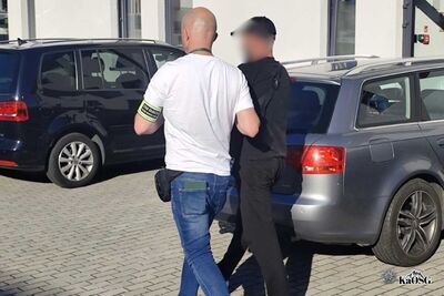 na zdjęciu jeden mężczyzna w ubraniu cywilnym i opaską na ramieniu z napisem straż graniczna prowadzi drugiego mężczyznę - prawdopodobnie osobę zatrzymają - w tle widoczny parking i samochody