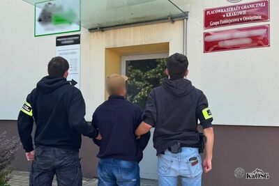 Na zdjęciu dwóch mężczyzn w cywilnych ubraniach i opaskach na ramieniu z napisem Straż Graniczna prowadzą pod rękę innego mężczyznę ubranego w jeansy oraz czarną bluzę i czapkę z daszkiem. Jest to najprawdopodobniej zatrzymany cudzoziemiec. W tle budynek z czerwoną tablicą z napisem Placówka Straży Granicznej w Krakowie - Grupa Zamiejscowa w Oświęcimiu,