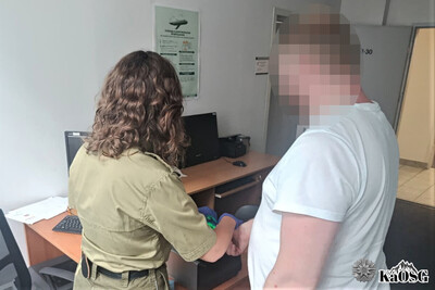 zatrzymany obywatel ukrainy stoi przy urządzeniu do daktyloskopii. funkcjonariuszka pobiera odciski palców zatrzymanemu cudzoziemcowi
